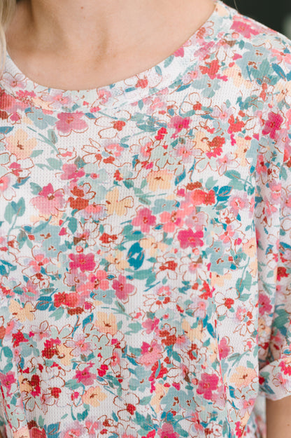 Close-up of a person wearing a short-sleeve top with a dense floral print in pink, coral, teal, and peach on a white base.