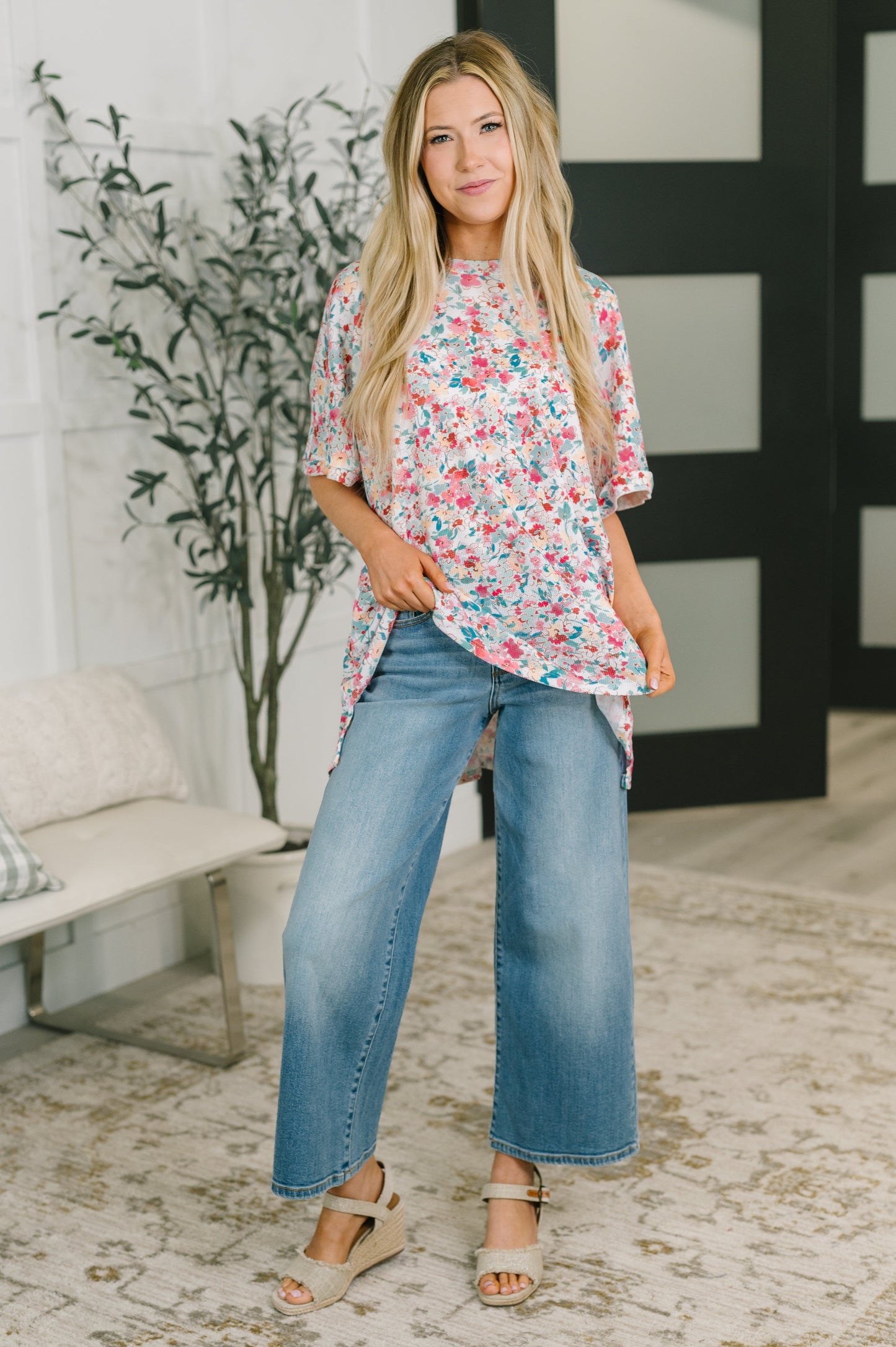 A woman modeling a short-sleeve floral top with blue wide-leg jeans and beige wedge sandals, standing in a bright indoor room with a bench, plant, and patterned rug.