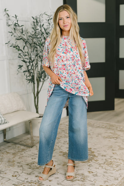 A woman modeling a short-sleeve floral top with blue wide-leg jeans and beige wedge sandals, standing in a bright indoor room with a bench, plant, and patterned rug.