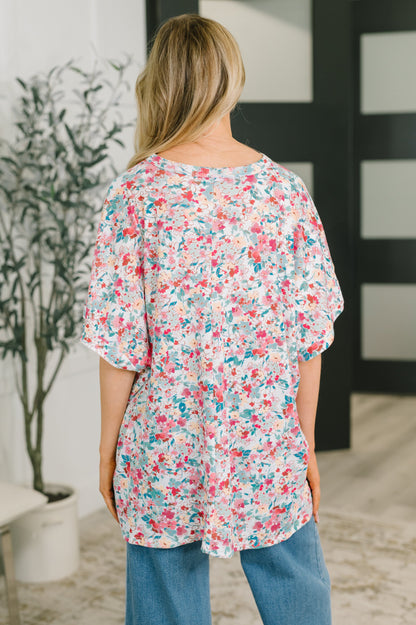 A woman viewed from behind wearing a loose short-sleeve floral top with pink, red, and blue flowers on a white background, paired with blue jeans in an indoor setting.