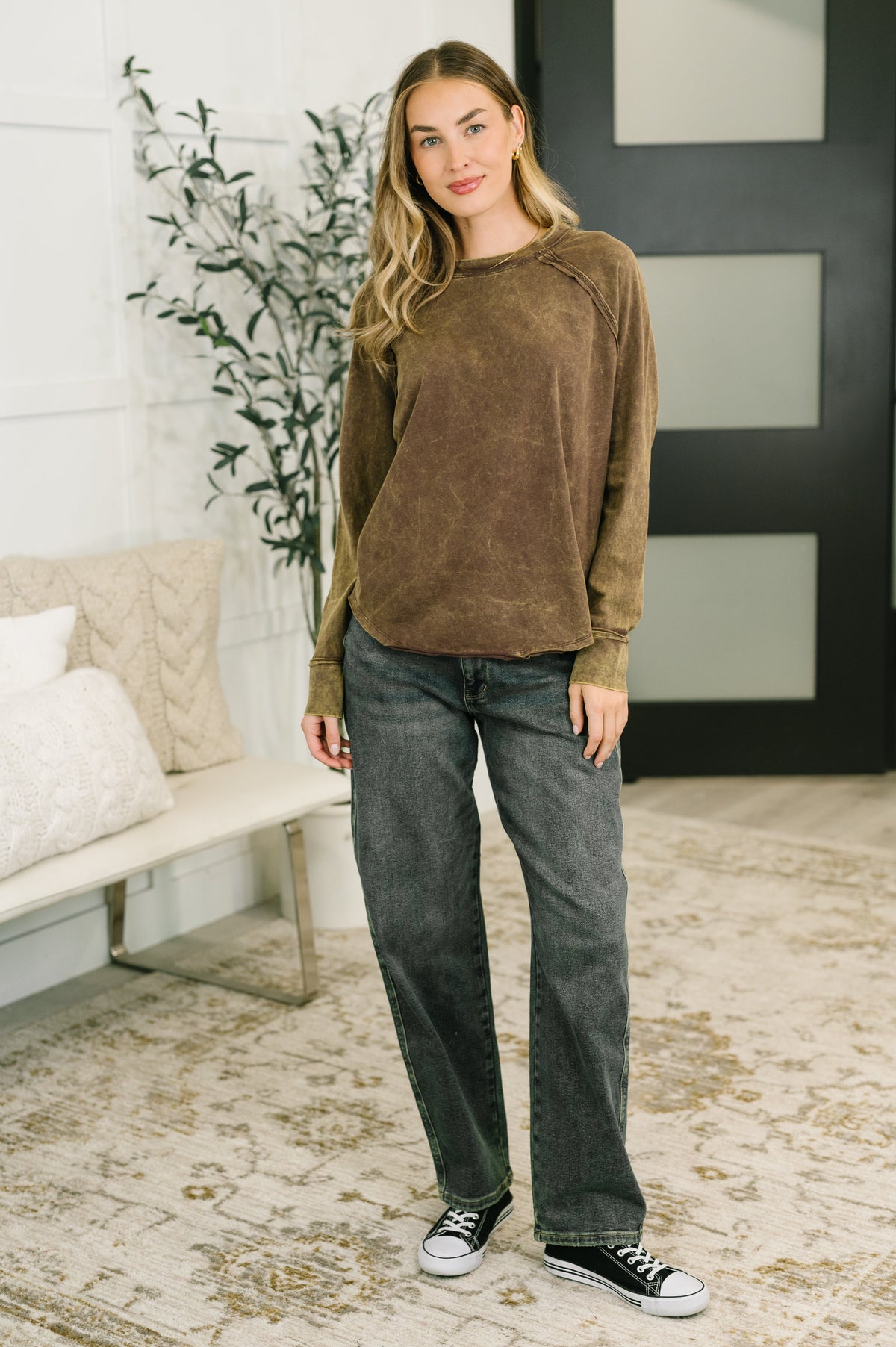 A woman modeling a brown long-sleeve crewneck top and dark gray jeans in a modern, neutral-toned living room setting.