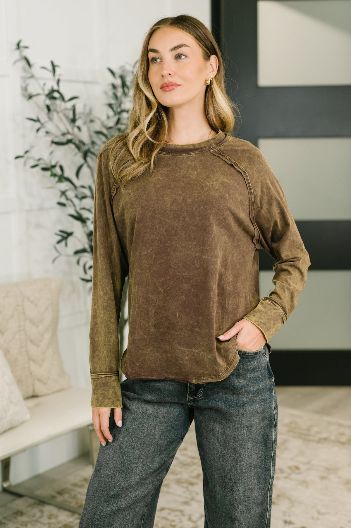 A woman modeling a brown mineral-wash long-sleeve top with raglan sleeves, paired with dark gray jeans, in a modern indoor setting.