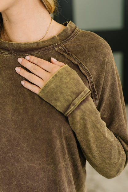 A close-up of a person wearing a brown long-sleeve crewneck top with a washed, textured finish. The person's hand rests on their chest, and a delicate gold necklace is visible.
