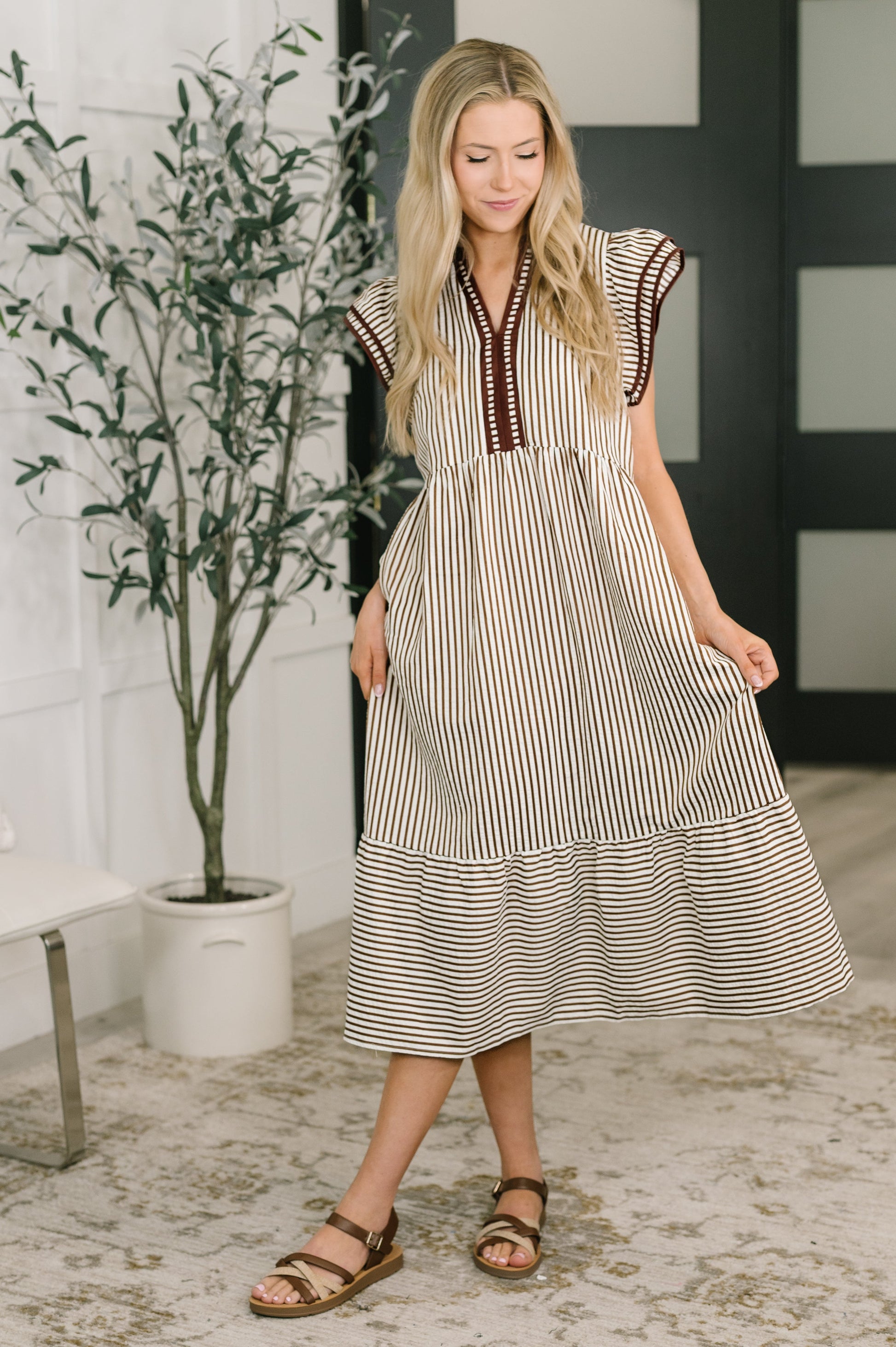 Woman wearing a striped dress standing in a room with a plant and bench.