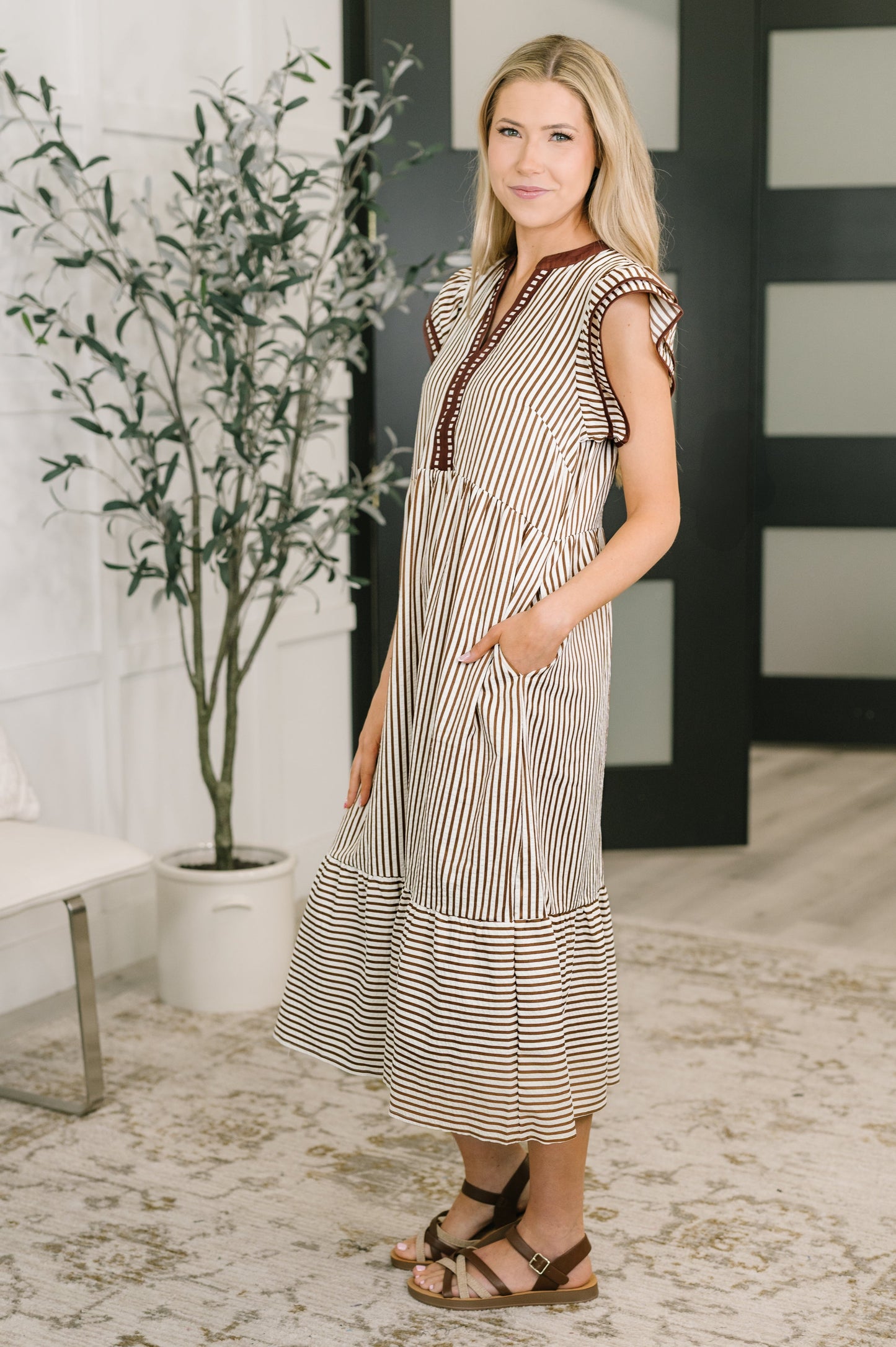 Woman wearing a striped dress standing in a room with a plant and bench.
