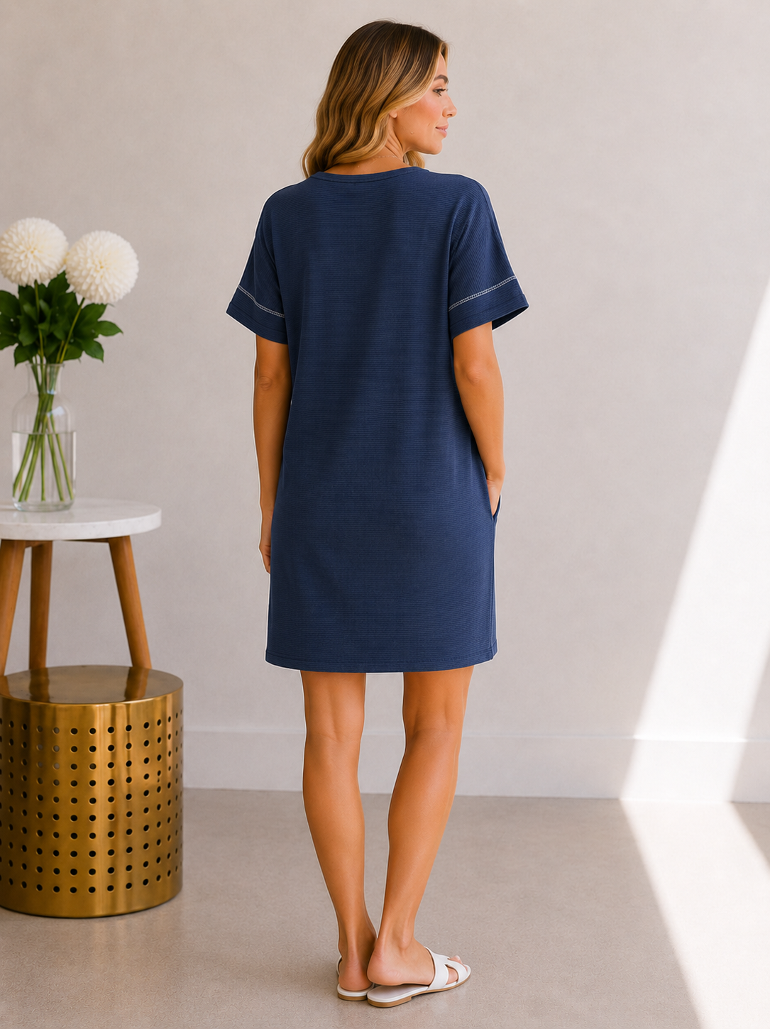 Woman wearing a navy blue dress standing in a minimalistic room with a vase of flowers on a table.