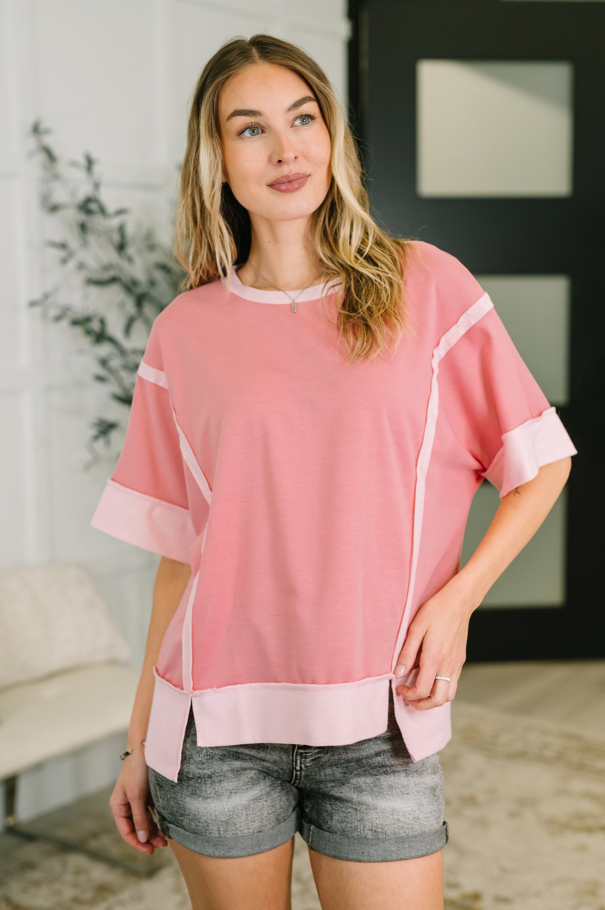Woman wearing a pink top with white accents and denim shorts indoors.