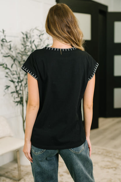 A woman viewed from behind wearing a black short-sleeve top with white contrast stitching along the neckline and sleeve hems, paired with blue jeans in an indoor setting.