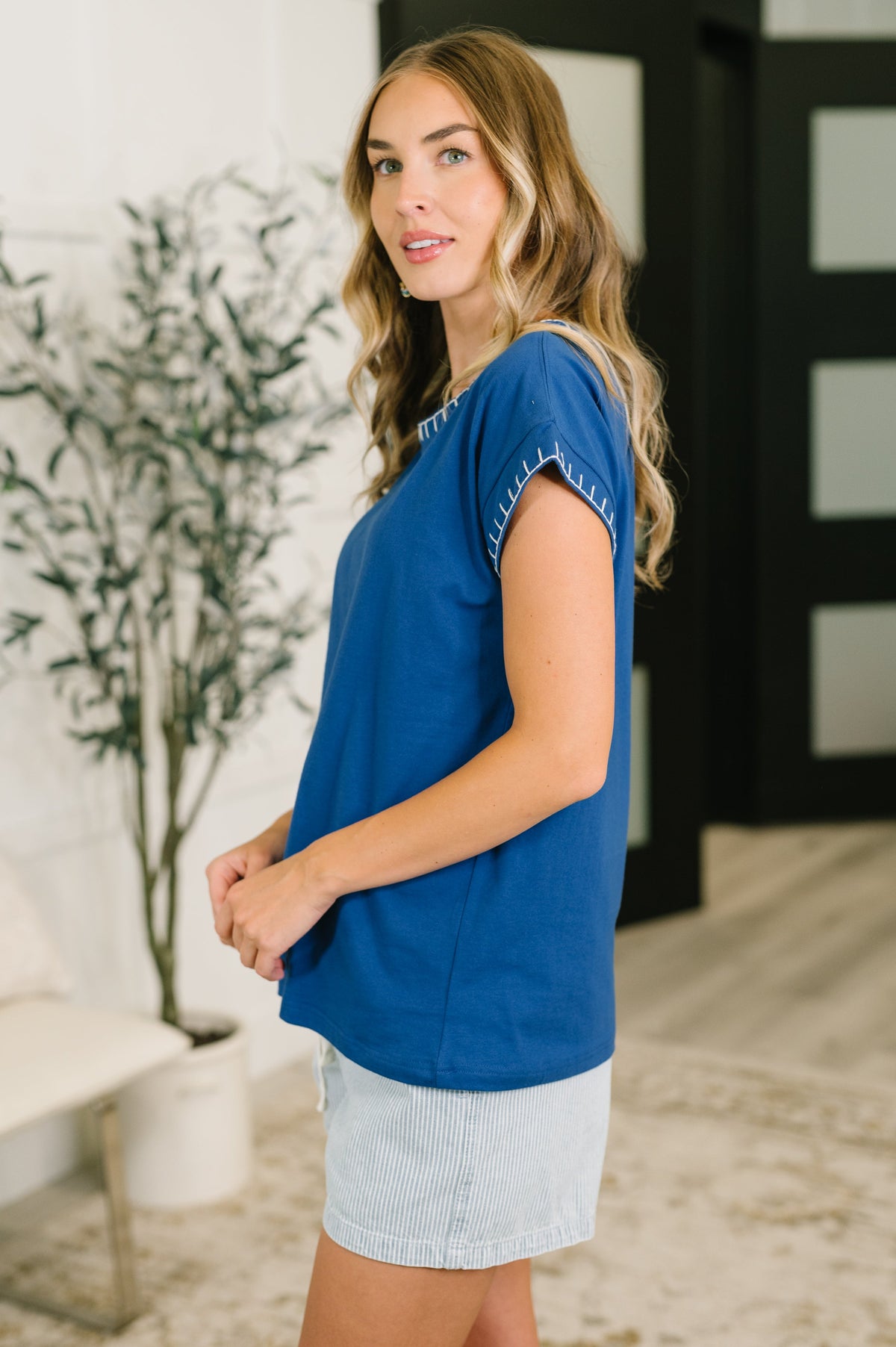 A woman modeling a royal blue short-sleeve top with decorative white trim along the sleeve edges, paired with a light denim skirt in a modern indoor setting.