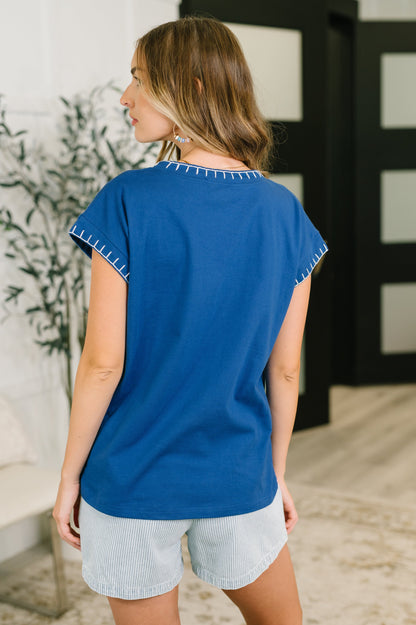 A woman shown from the back wearing a royal blue short-sleeve top with white contrast stitching along the collar and sleeve hems, paired with light gray shorts in an indoor setting.