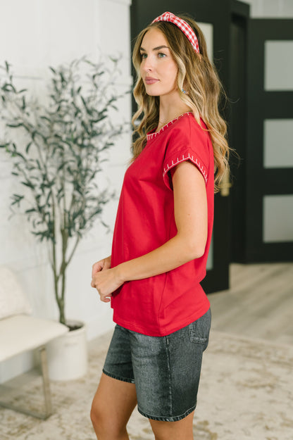 A woman modeling a bright red short-sleeve top with decorative white stitching along the sleeve hems, paired with denim shorts and a red-and-white gingham headband in a modern indoor setting.