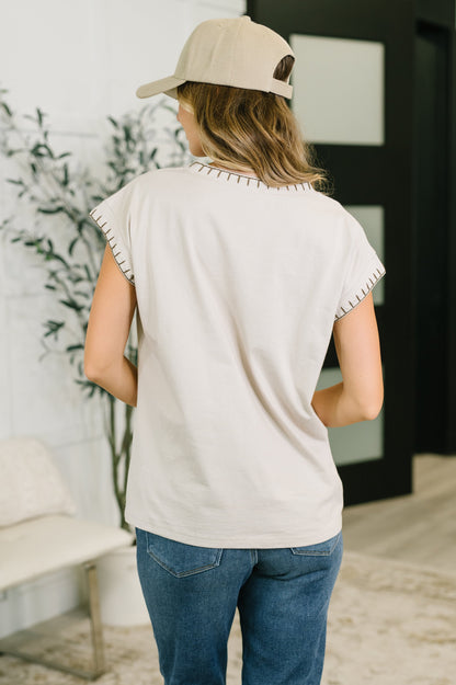 A woman viewed from behind wearing a light beige short-sleeve t-shirt with scalloped trim, blue jeans, and a beige baseball cap in a modern indoor setting.