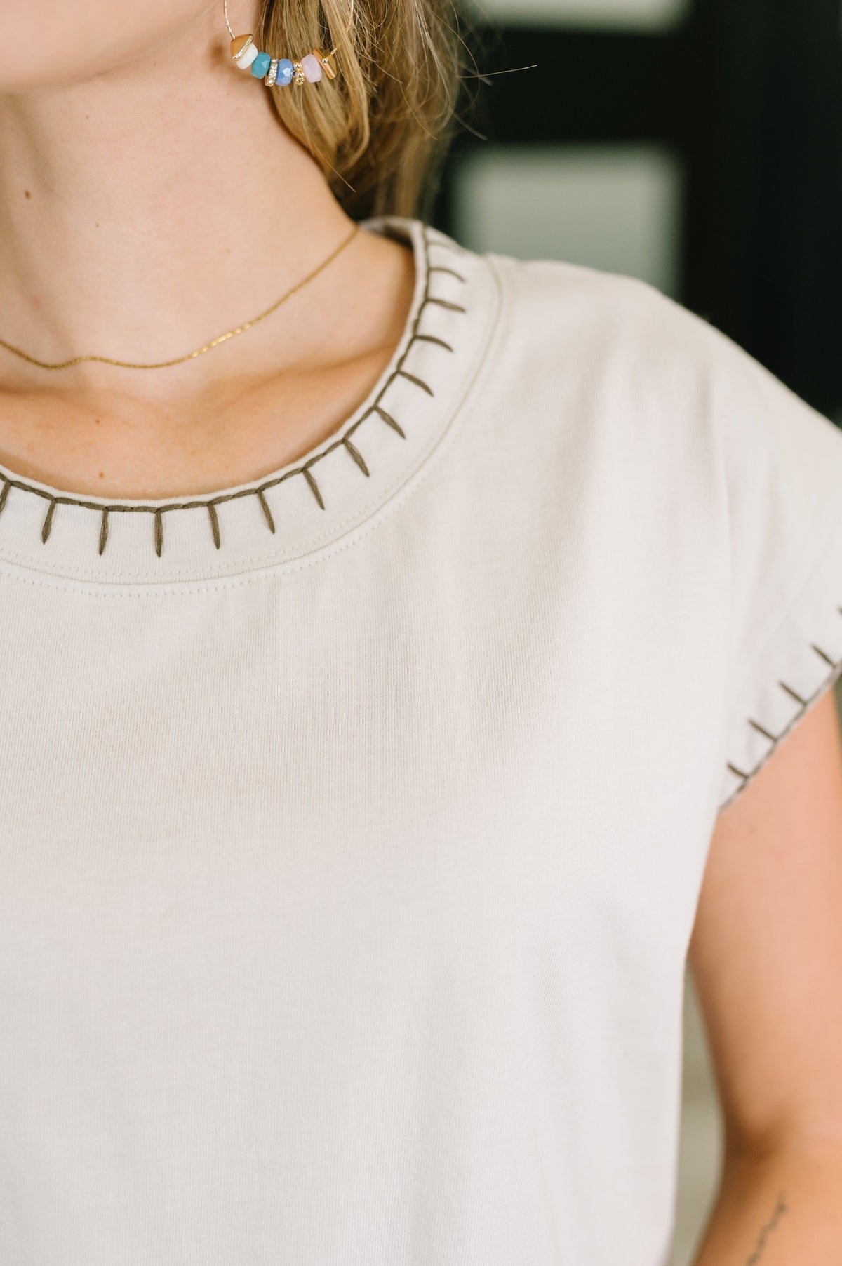 Close-up of a person wearing a light beige or off-white short-sleeve top with decorative dark stitched details around the neckline and sleeve edges.