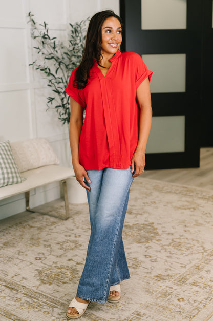 Woman wearing a red top and blue jeans standing in a room with a plant and bench.