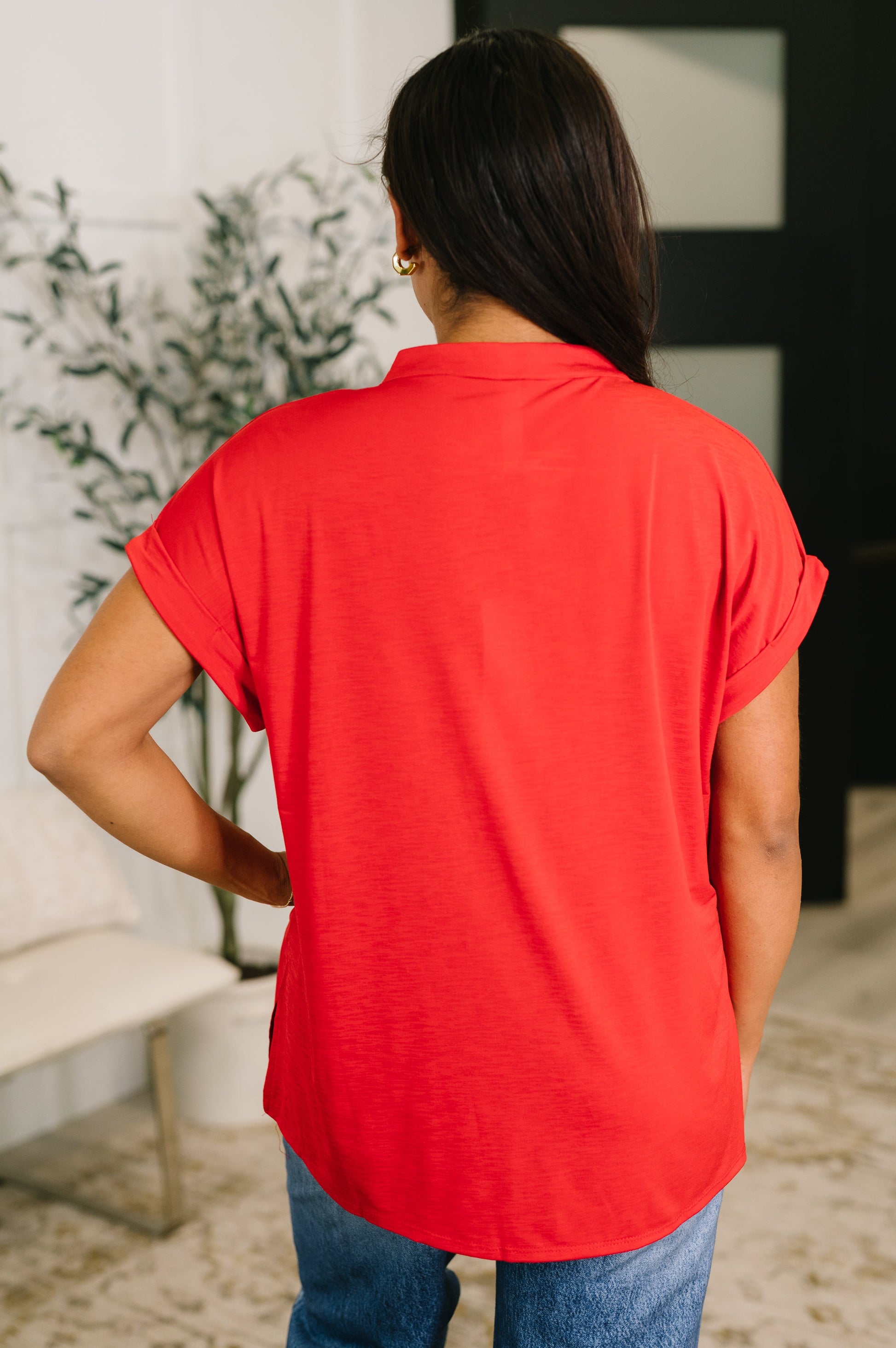 Person wearing a red shirt and blue jeans standing in a room with a plant and furniture.