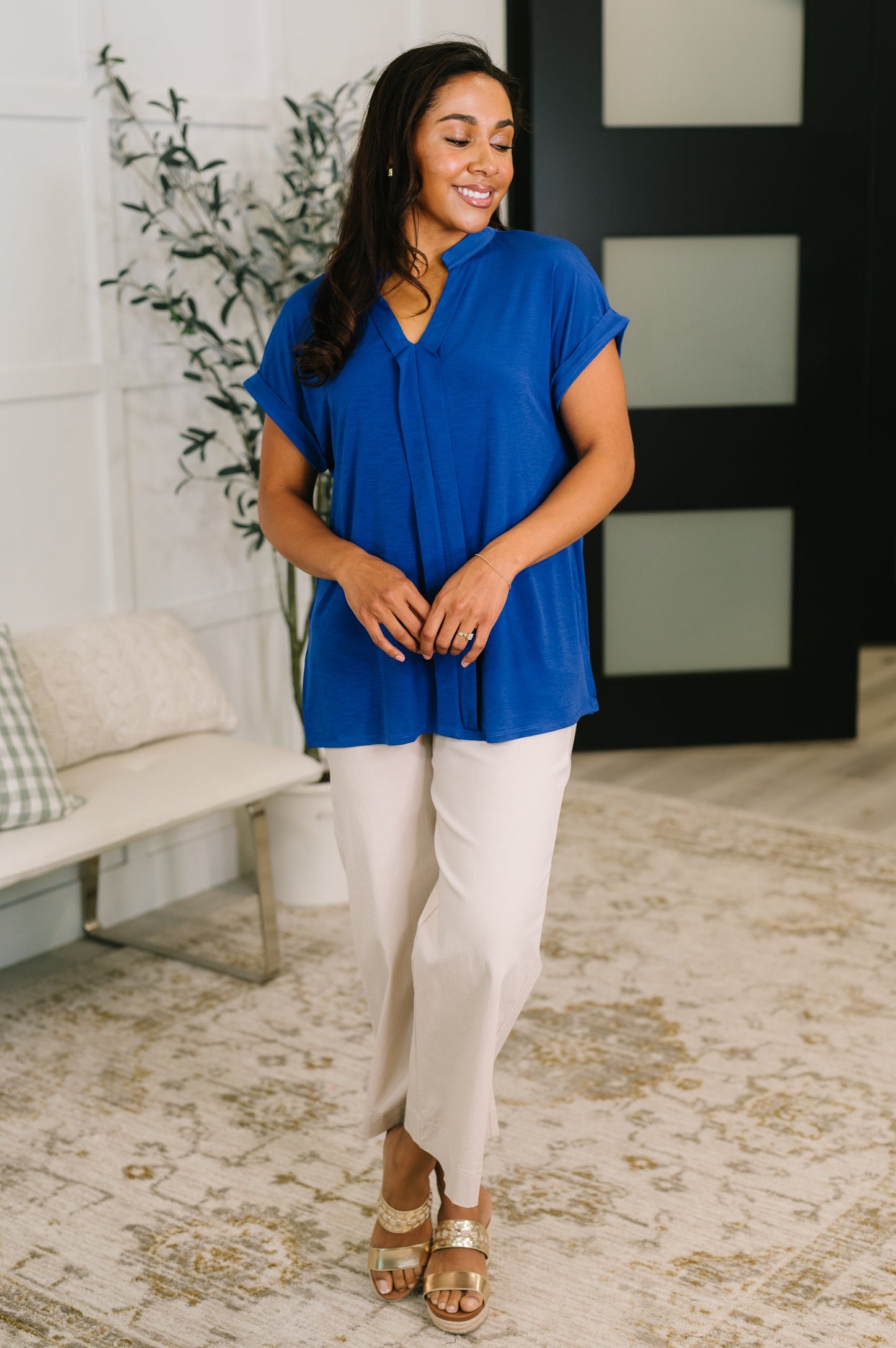 Woman wearing a blue top and beige pants standing in a room with a plant and bench.