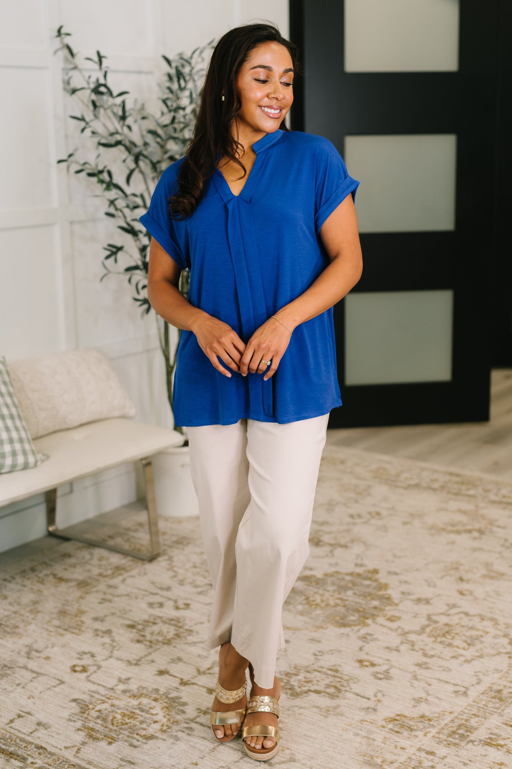 Woman wearing a blue top and beige pants standing in a room with a plant and bench.