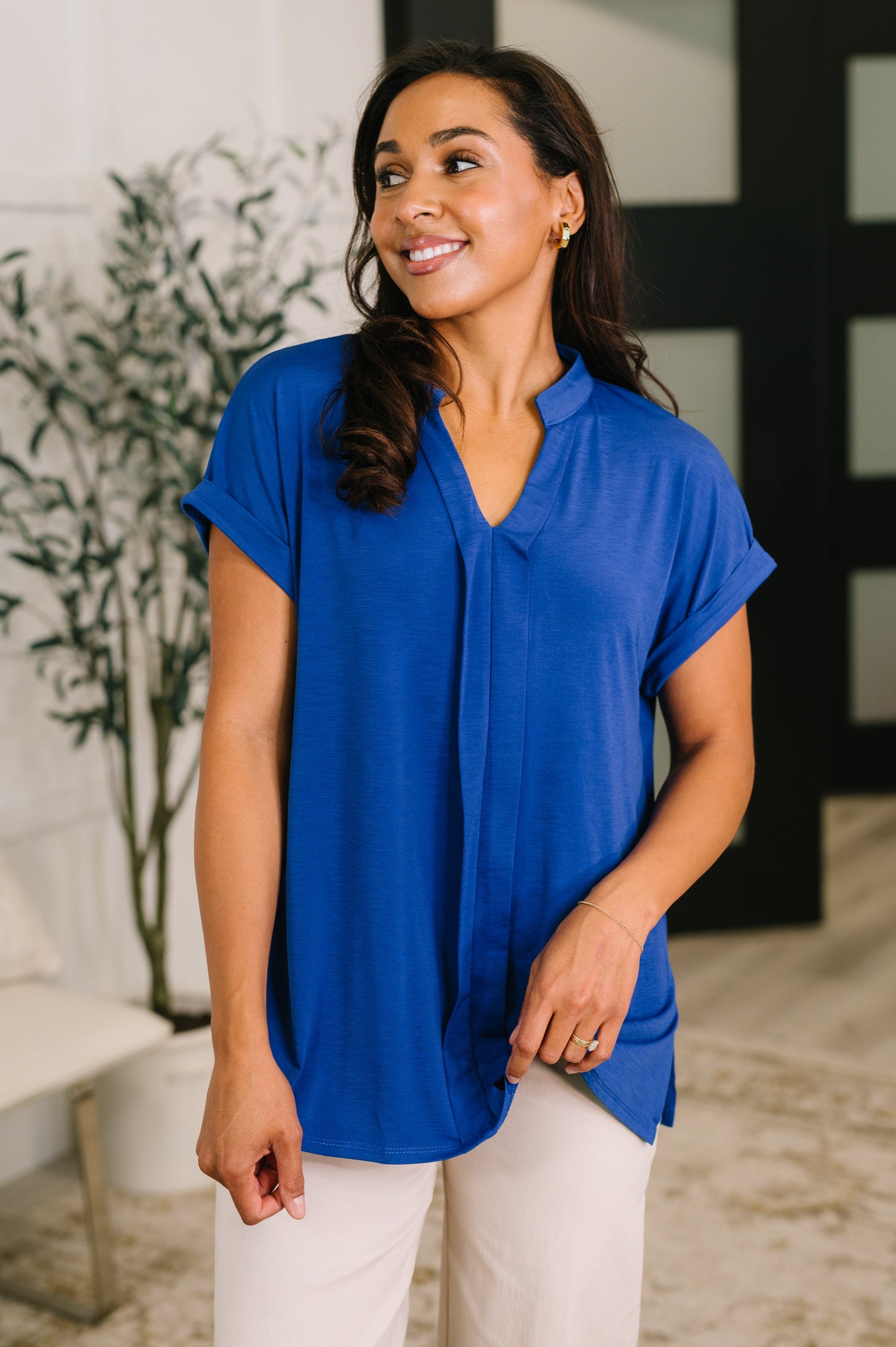 Woman wearing a blue blouse standing indoors with a plant and bench in the background