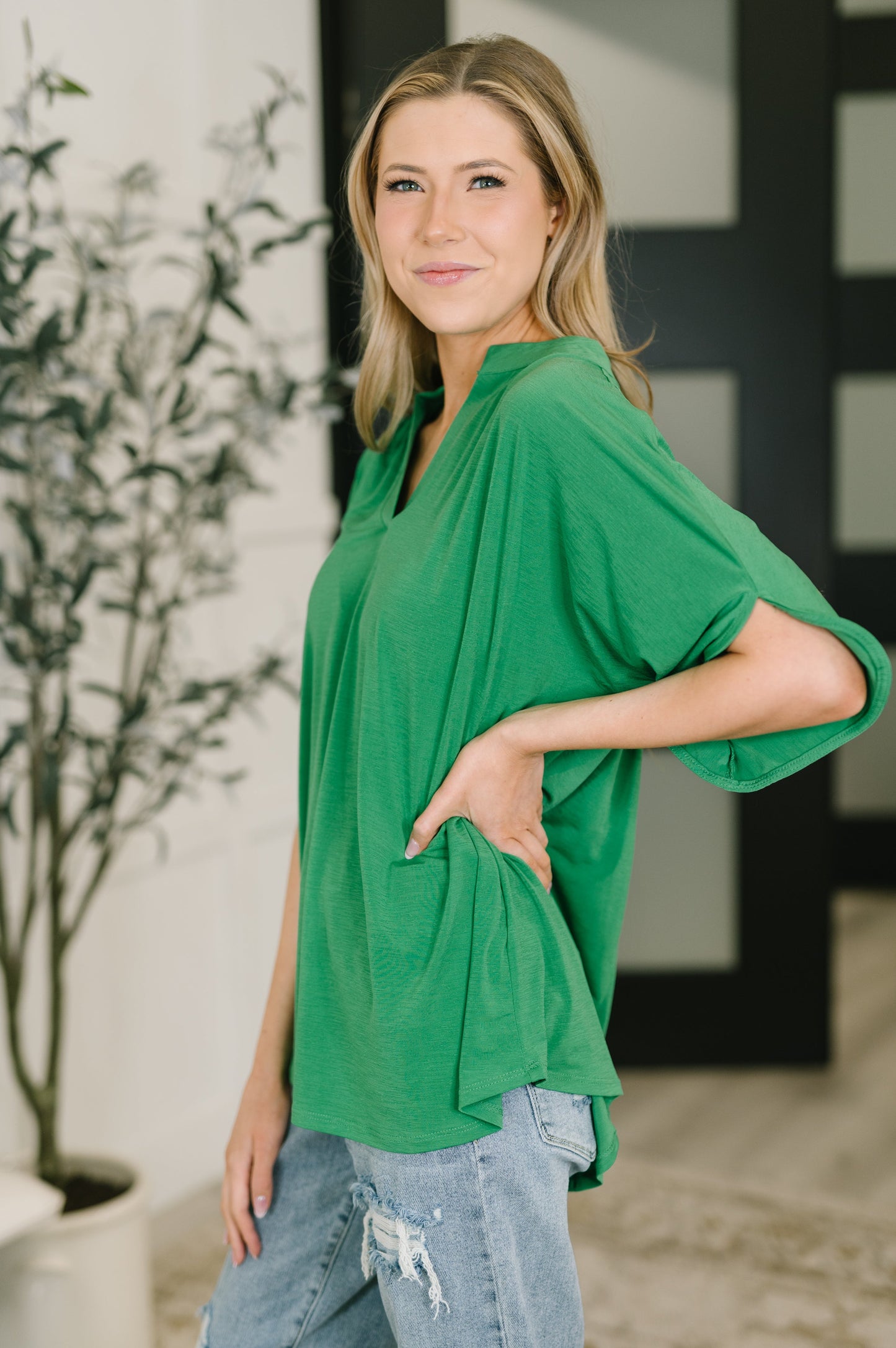 Woman wearing a green top and blue jeans standing indoors with a plant in the background