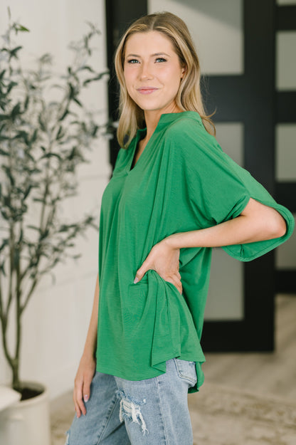 Woman wearing a green top and blue jeans standing indoors with a plant in the background