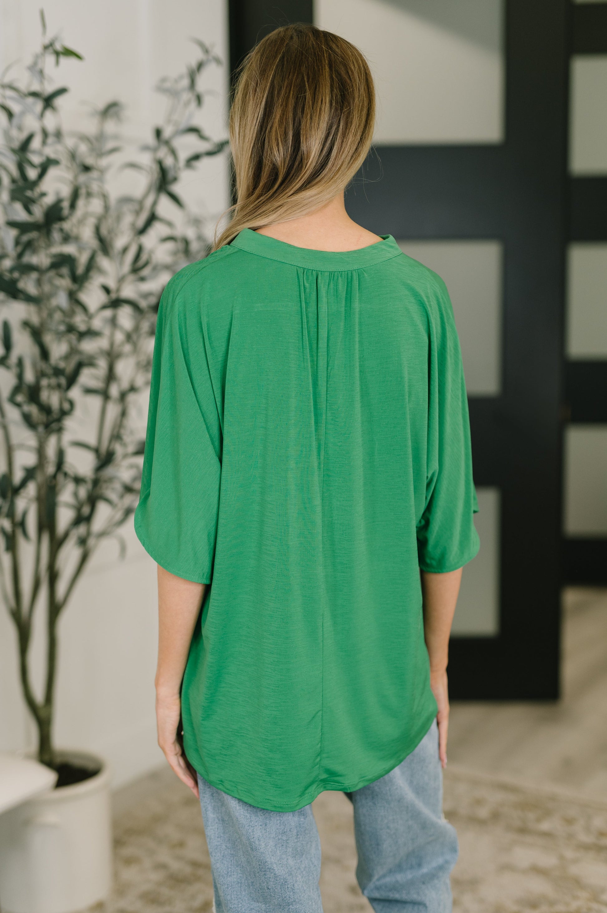 Person wearing a green top in an indoor setting with a plant and decorative wall.