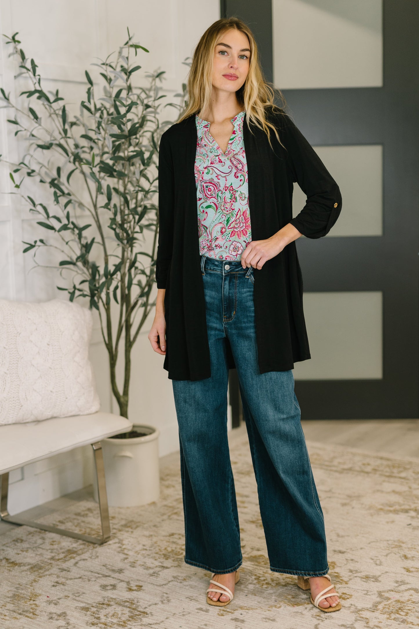 Woman wearing a black cardigan, floral top, and blue jeans in a living room setting.