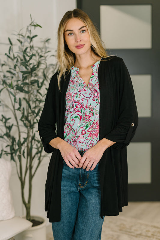 Woman wearing a black cardigan over a patterned shirt with blue jeans, standing indoors.