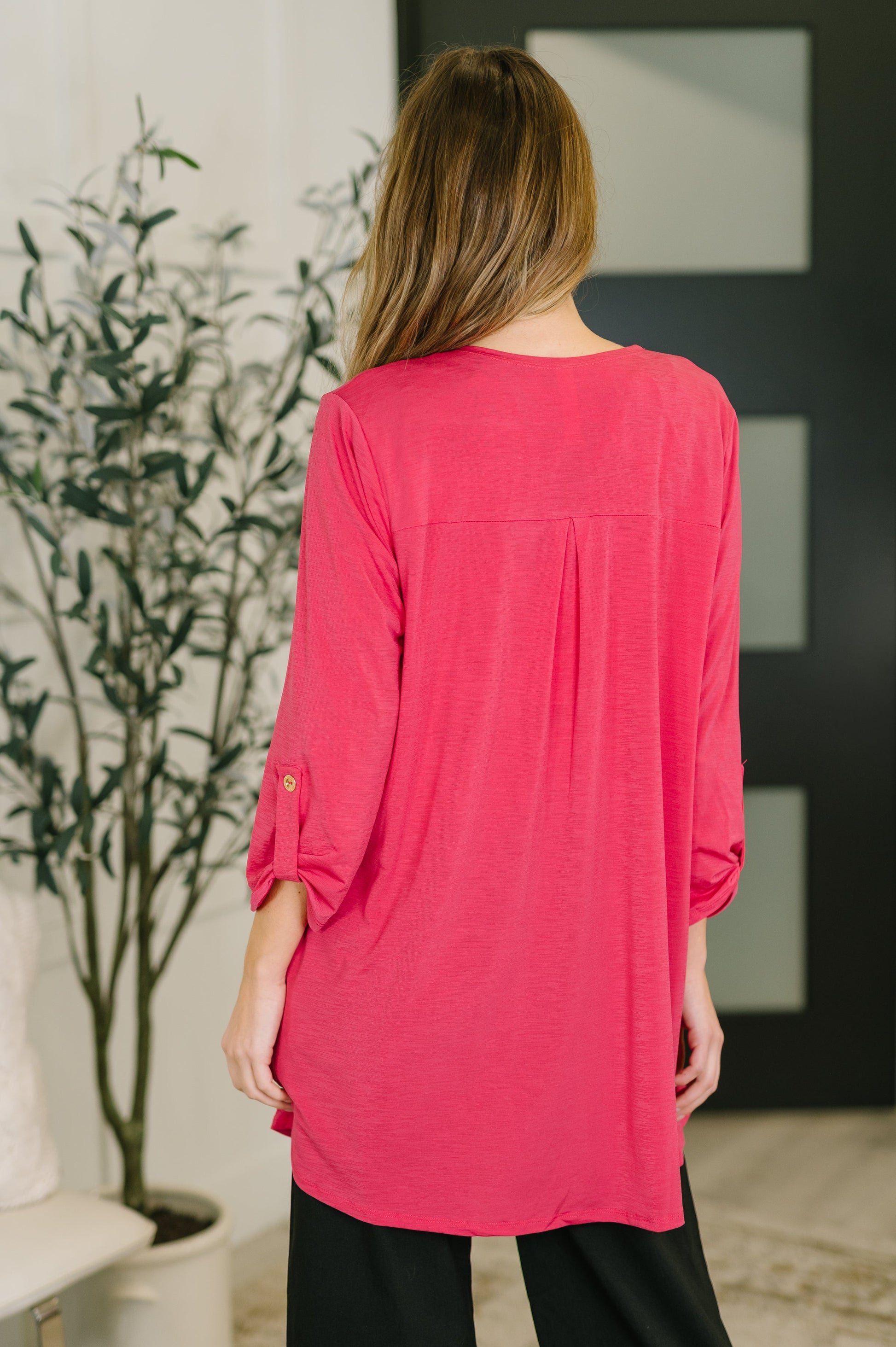 Person wearing a pink long-sleeve top in an indoor setting with a plant and wall decor.
