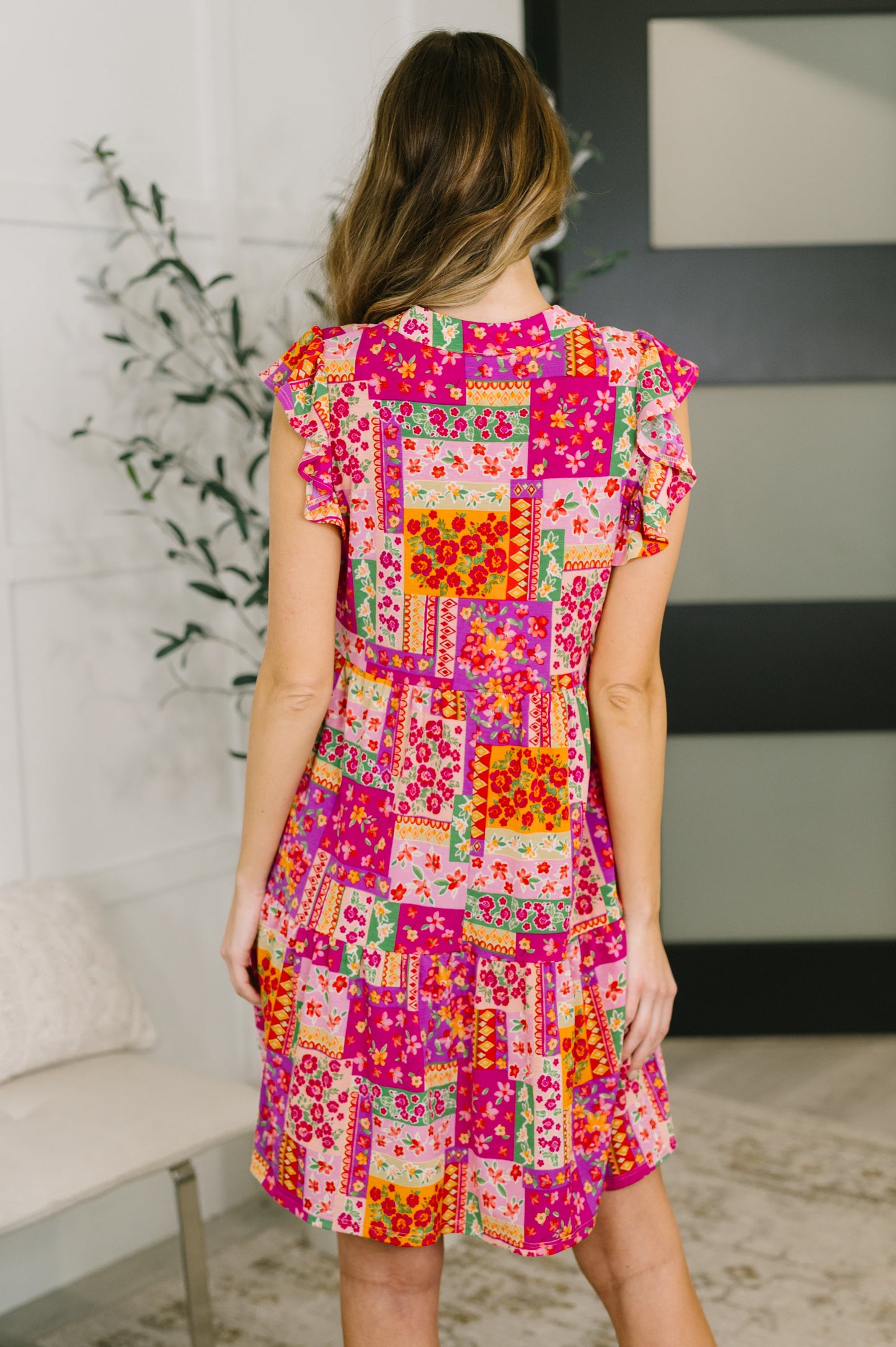 Woman wearing a colorful patchwork dress in a room with a plant and sofa.
