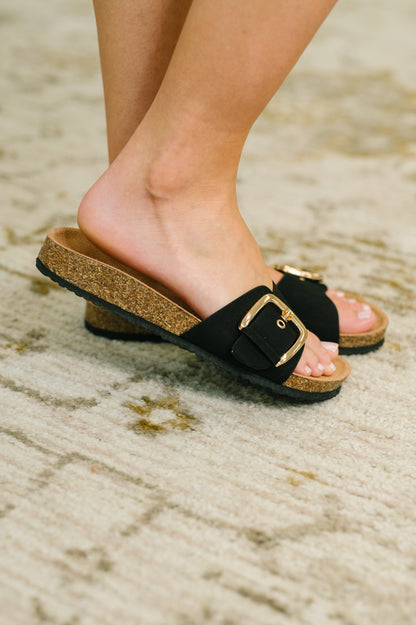Black sandals with gold buckles worn on a sandy surface
