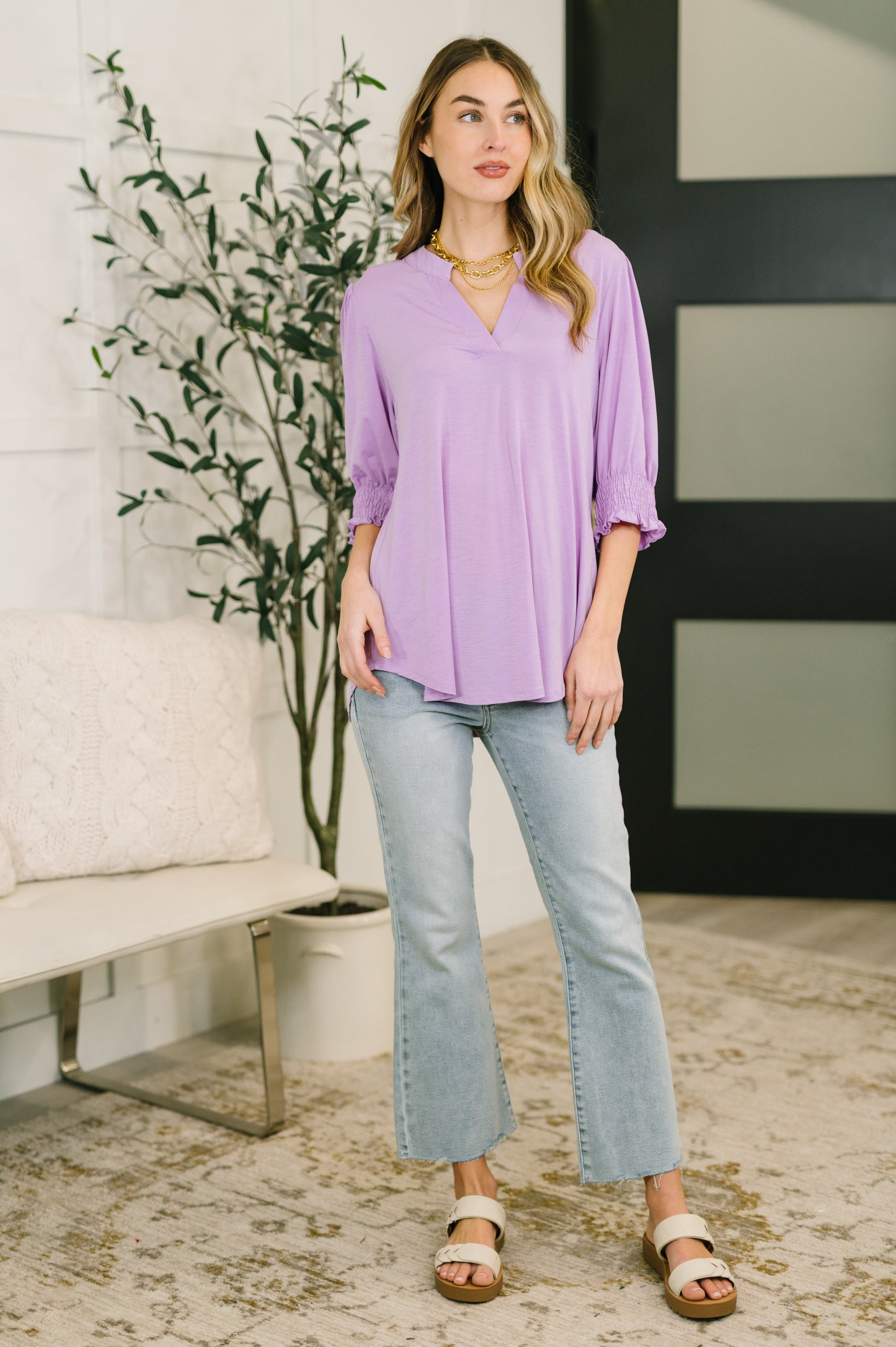 Woman wearing a purple blouse and light blue jeans standing in a room with a plant and furniture.