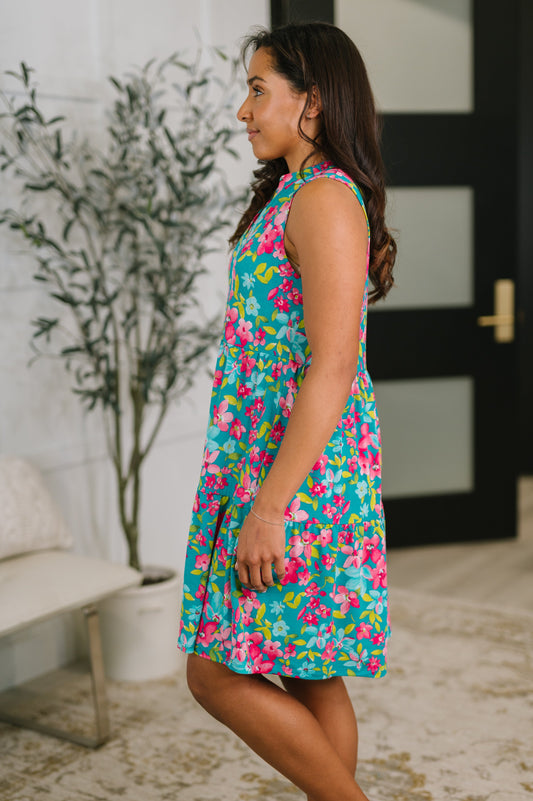 Woman wearing a colorful floral dress standing in a room with a plant and door in the background.