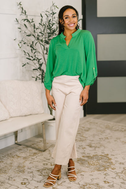 Woman wearing a green blouse and beige pants standing in a room with a plant and bench.