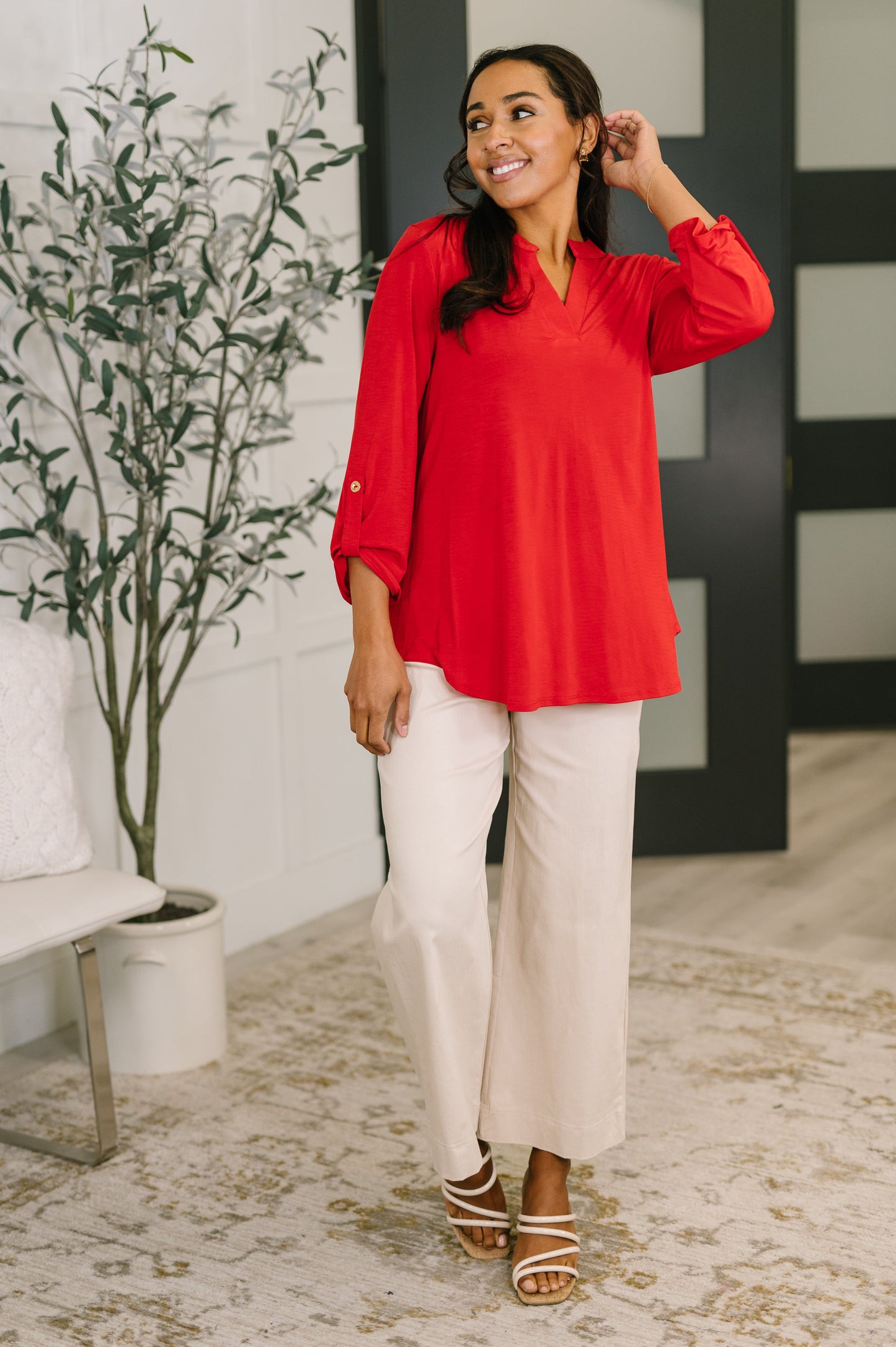 Woman wearing a red blouse and beige pants standing in a room with a plant and decorative rug.