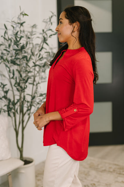Woman wearing a red blouse standing indoors with a plant and wall decor in the background