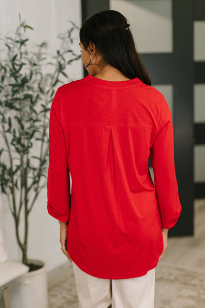 Person wearing a red long-sleeve top in an indoor setting with a plant and chair in the background.