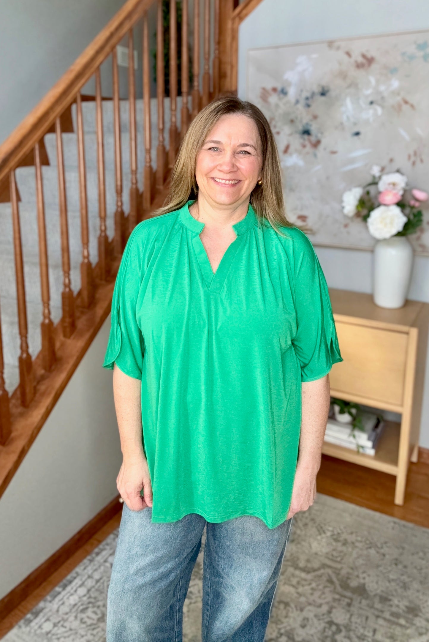 Woman wearing a green blouse and blue jeans standing in a home setting with a staircase and decorative items.