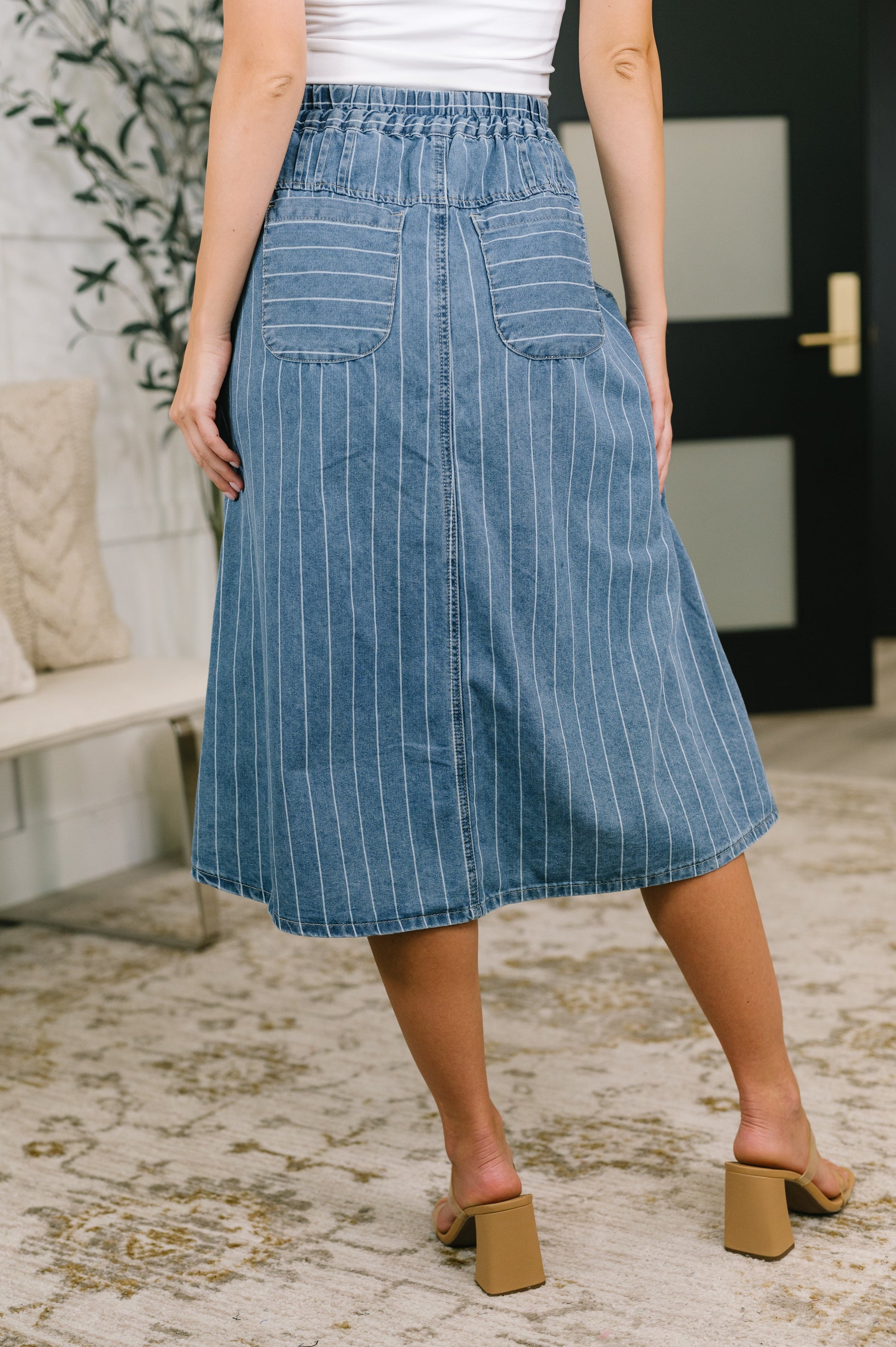A person wearing a blue denim midi skirt with thin white vertical pinstripes, shown from the back in an indoor setting with a patterned rug and a bench in the background.