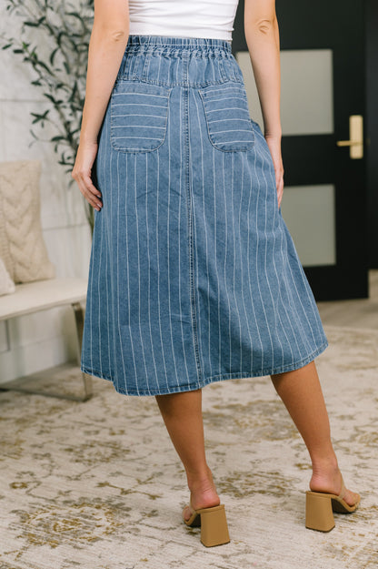 A person wearing a blue denim midi skirt with thin white vertical pinstripes, shown from the back in an indoor setting with a patterned rug and a bench in the background.