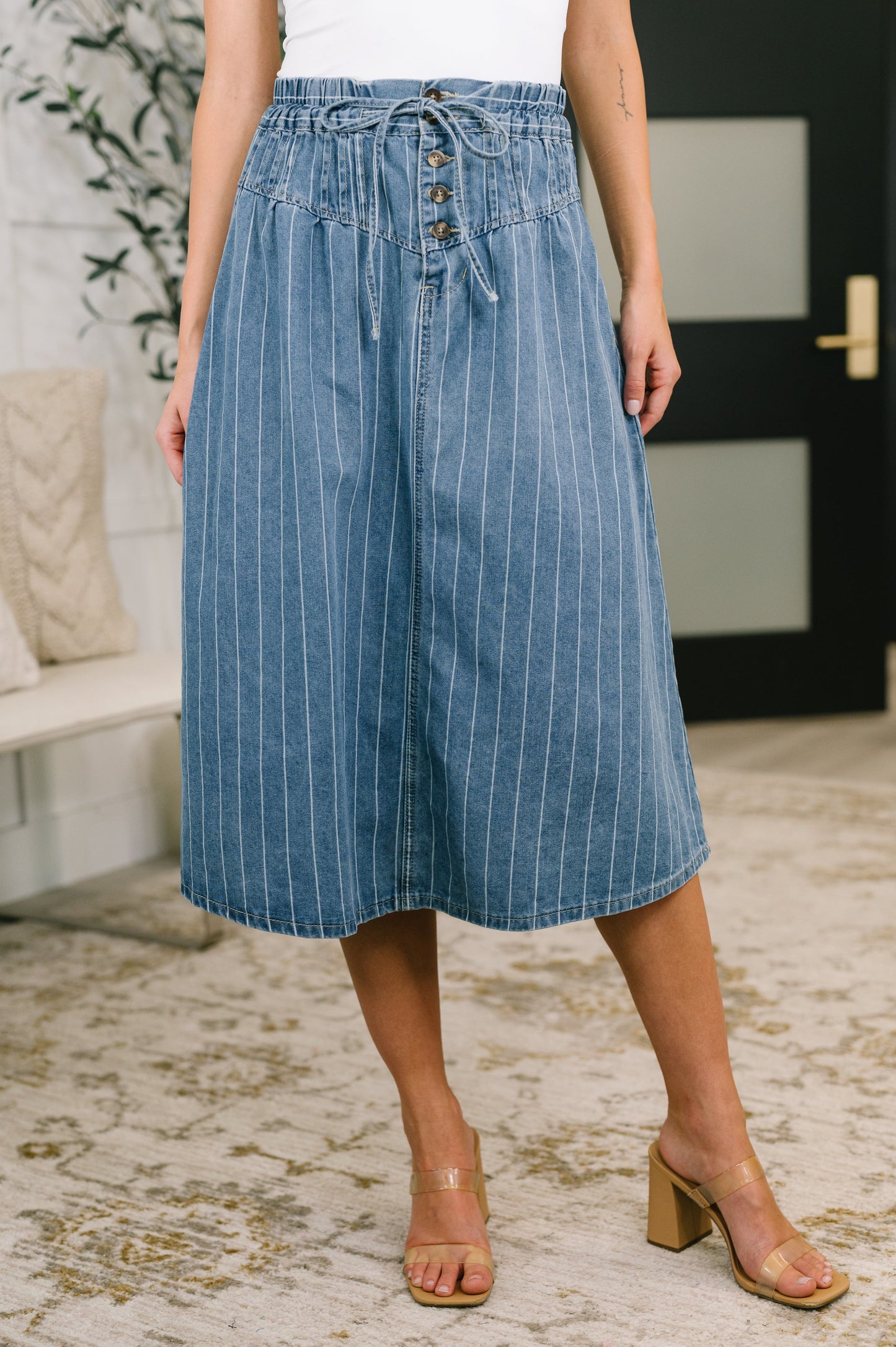 A model wearing a blue denim midi skirt with thin white vertical pinstripes, a drawstring waist, and button details down the front, paired with beige block-heel sandals in an indoor setting.
