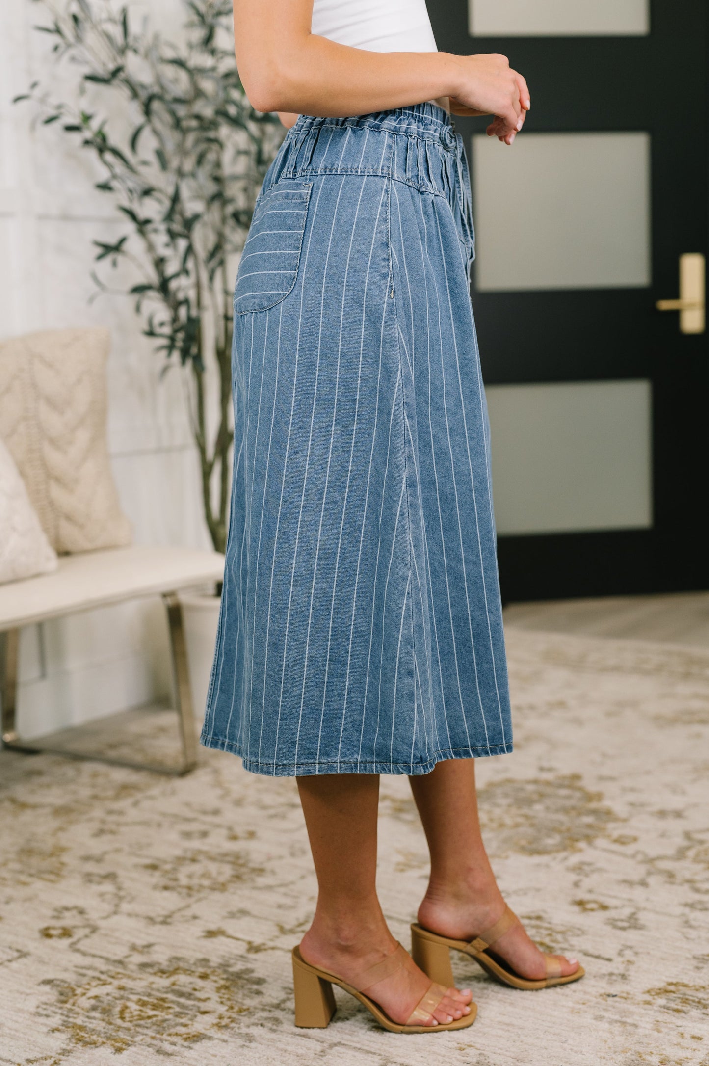 A person wearing a blue denim midi skirt with thin white vertical pinstripes, paired with a white top and tan block-heel slide sandals, standing in a modern indoor setting.
