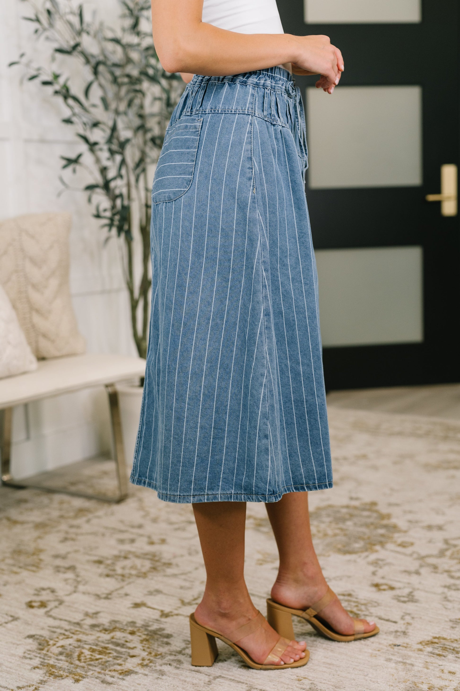 A person wearing a blue denim midi skirt with thin white vertical pinstripes, paired with a white top and tan block-heel slide sandals, standing in a modern indoor setting.
