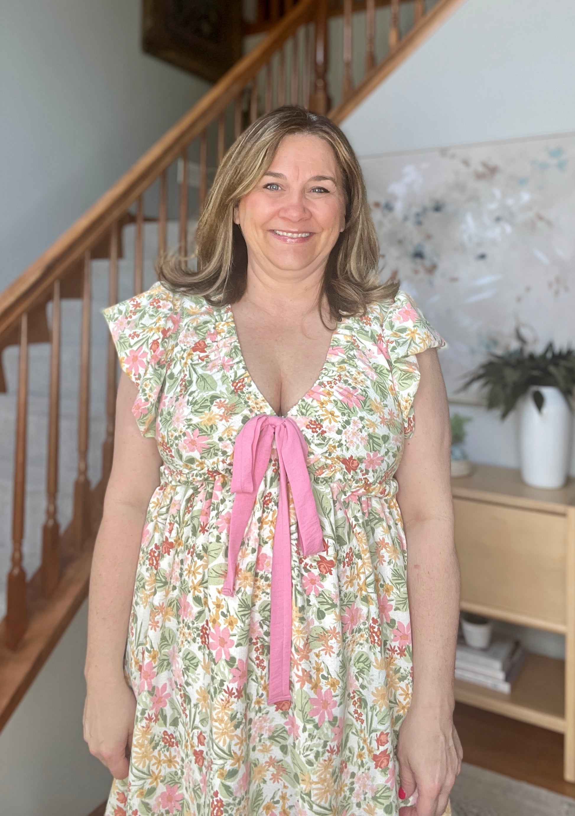Woman wearing a floral dress with a pink ribbon standing in a home setting.