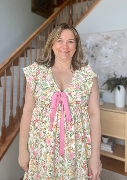 Woman wearing a floral dress with a pink ribbon standing in a home setting.