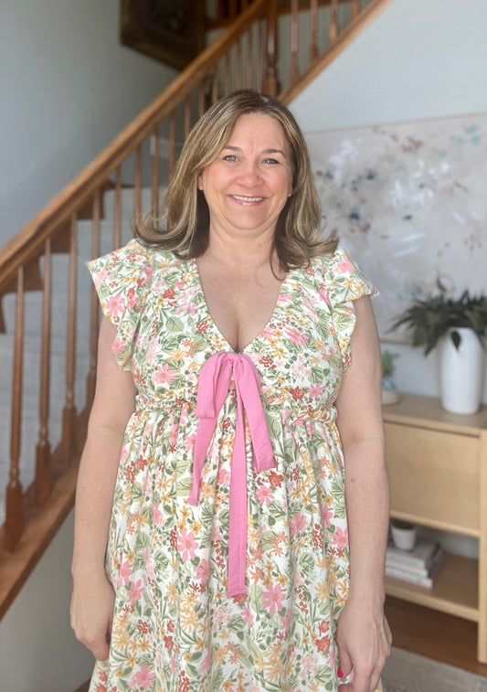 Woman wearing a floral dress with a pink ribbon standing in a home setting.