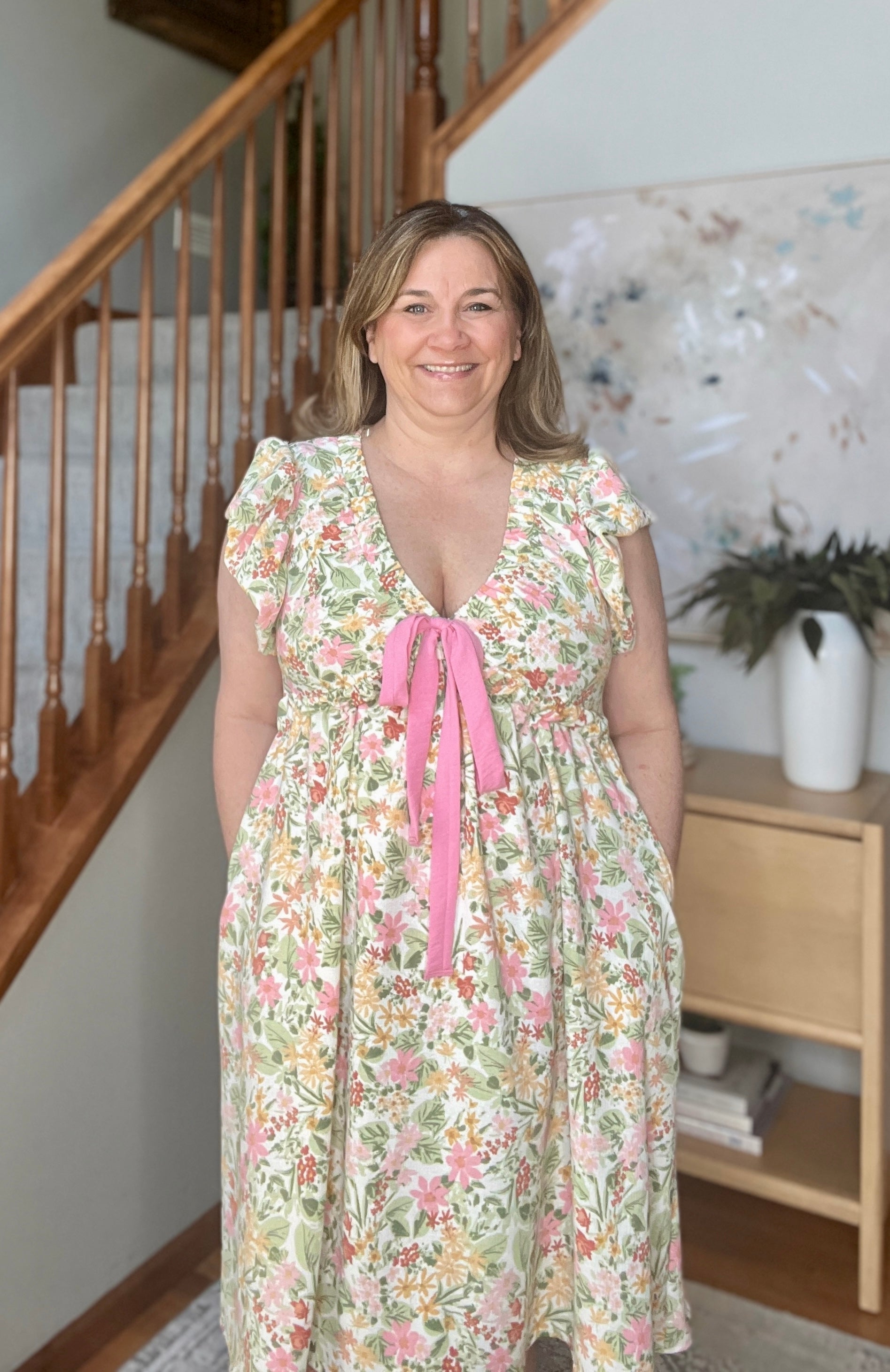 Woman wearing a v-neck floral dress with a pink ribbon standing in a home setting.