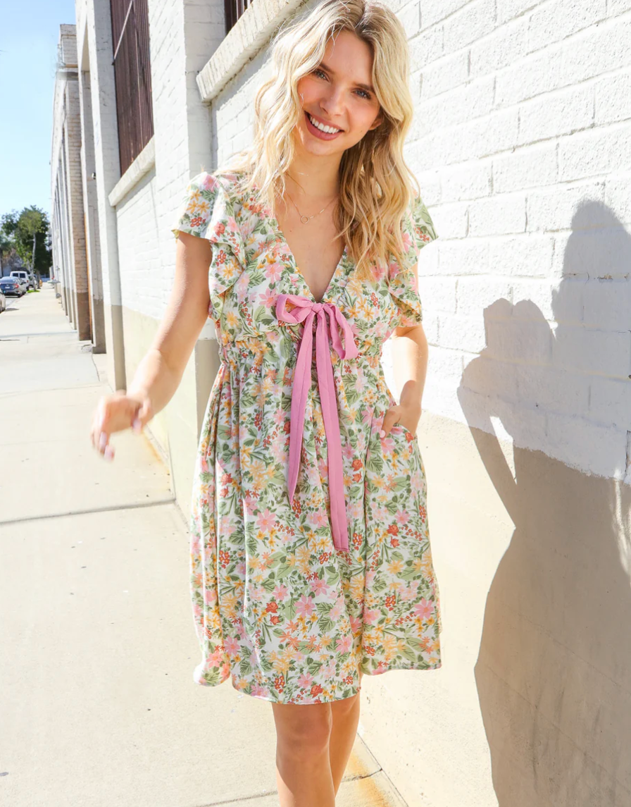 Woman wearing a floral dress with a pink ribbon standing against a white wall.