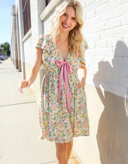 Woman wearing a floral dress with a pink ribbon standing against a white wall.