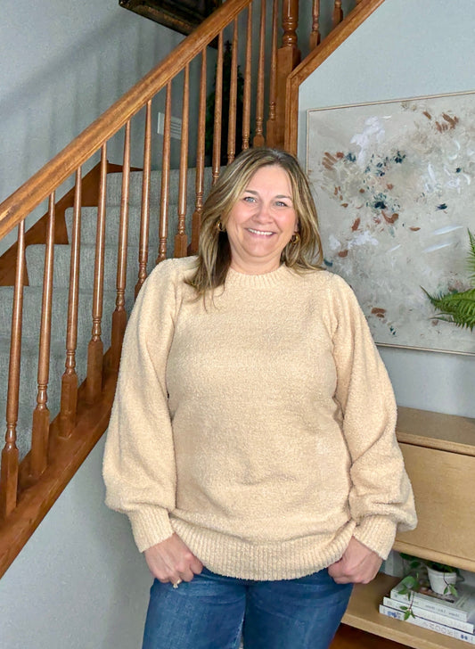Woman wearing a beige sweater standing in a home setting with a staircase and artwork in the background.