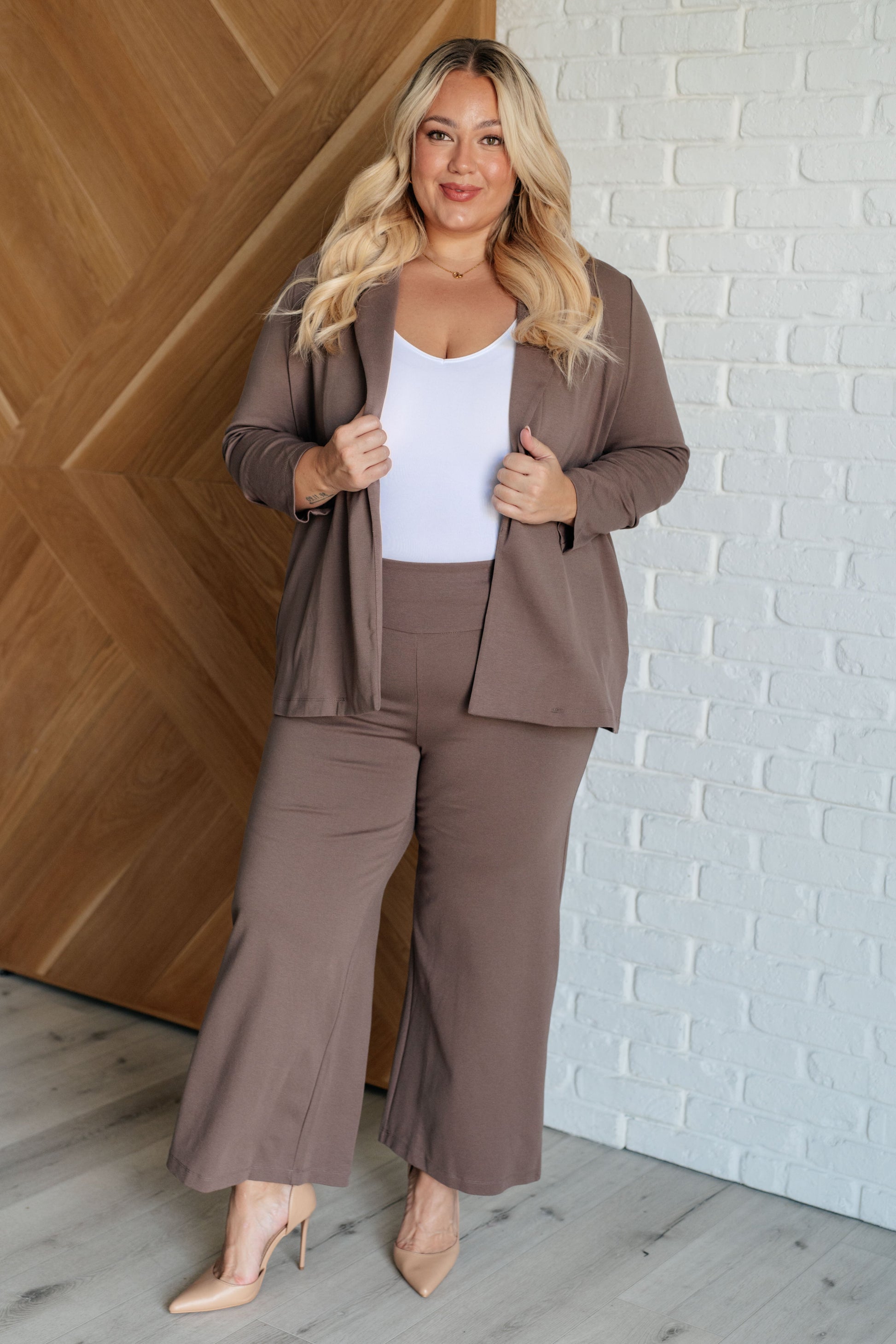 Woman in a brown suit standing against a white brick wall.